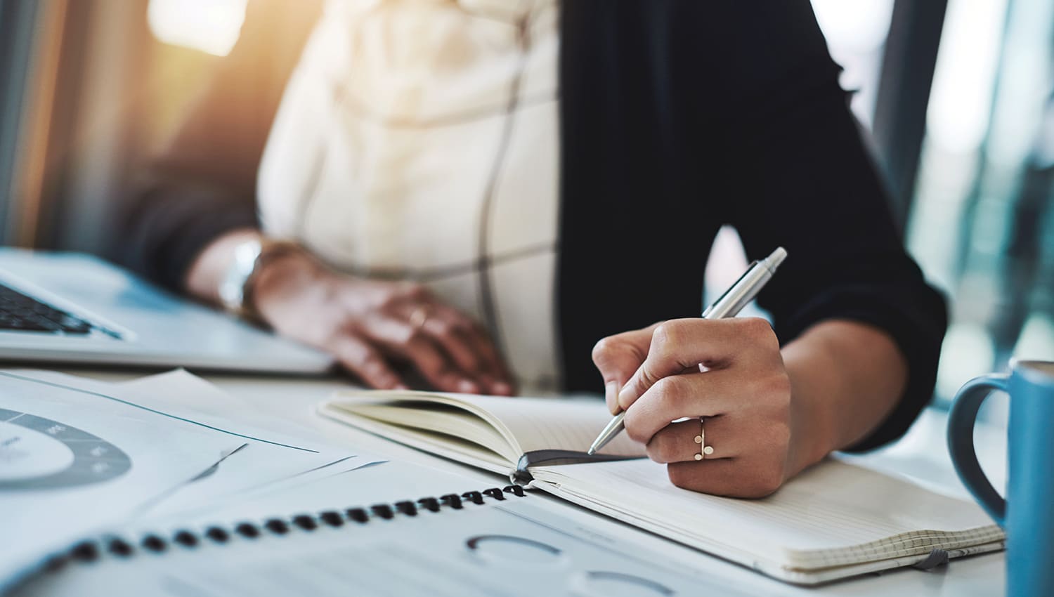 A woman wearing a black sweater holds a silver pen in her hand as she takes notes about her company's workers' compensation program in an empty notebook. laptop and stack of books